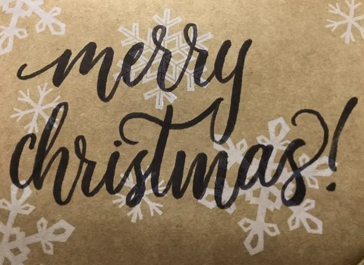 Merry Christmas! in black brush script on kraft paper with white snowflakes