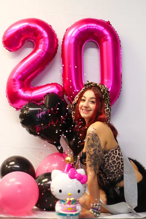 A woman posing beside pink 20 balloons and a Hello Kitty cake