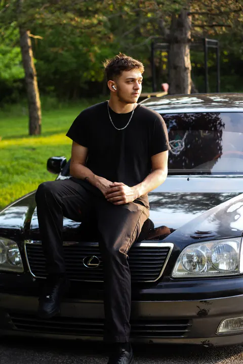 A young man sitting on the hood of a black Lexus