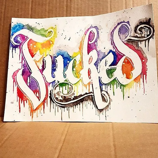 A swear word in rainbow watercolour blackletter with drips, on paper propped on cardboard